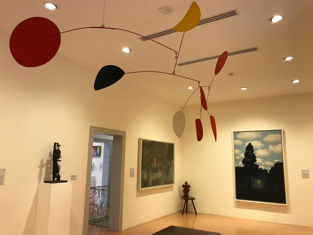 이동식 조각품이 걸려 있는 미술관, 벽에 걸린 그림, 출입구, 작은 조각상이 있는 받침대.