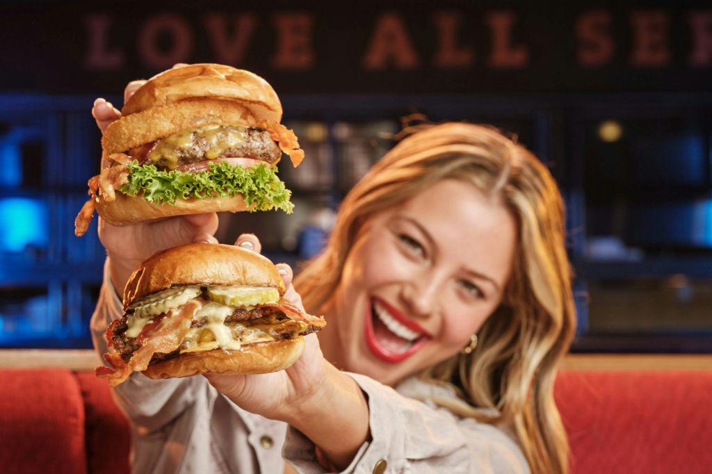 레스토랑을 배경으로 양손에 하나씩 햄버거 두 개를 들고 미소 짓는 사람.