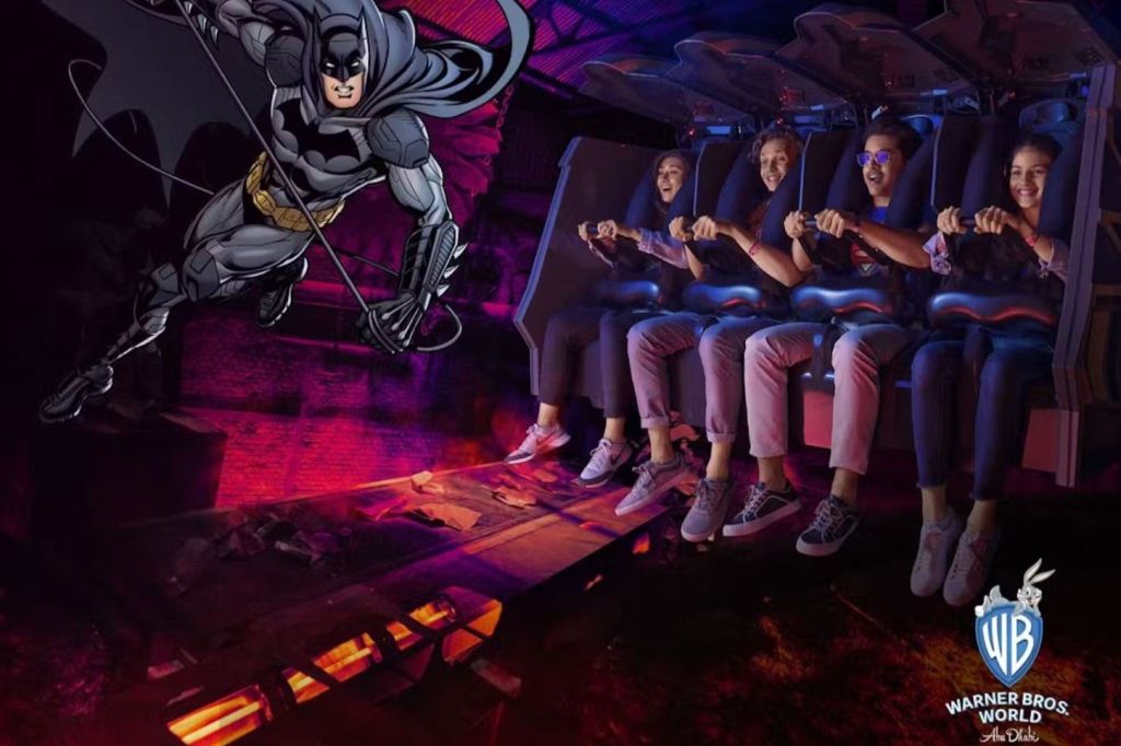 극적인 배경과 워너 브라더스 로고가 보이는 놀이 기구에 4명이 앉아 손잡이를 잡고 있습니다.