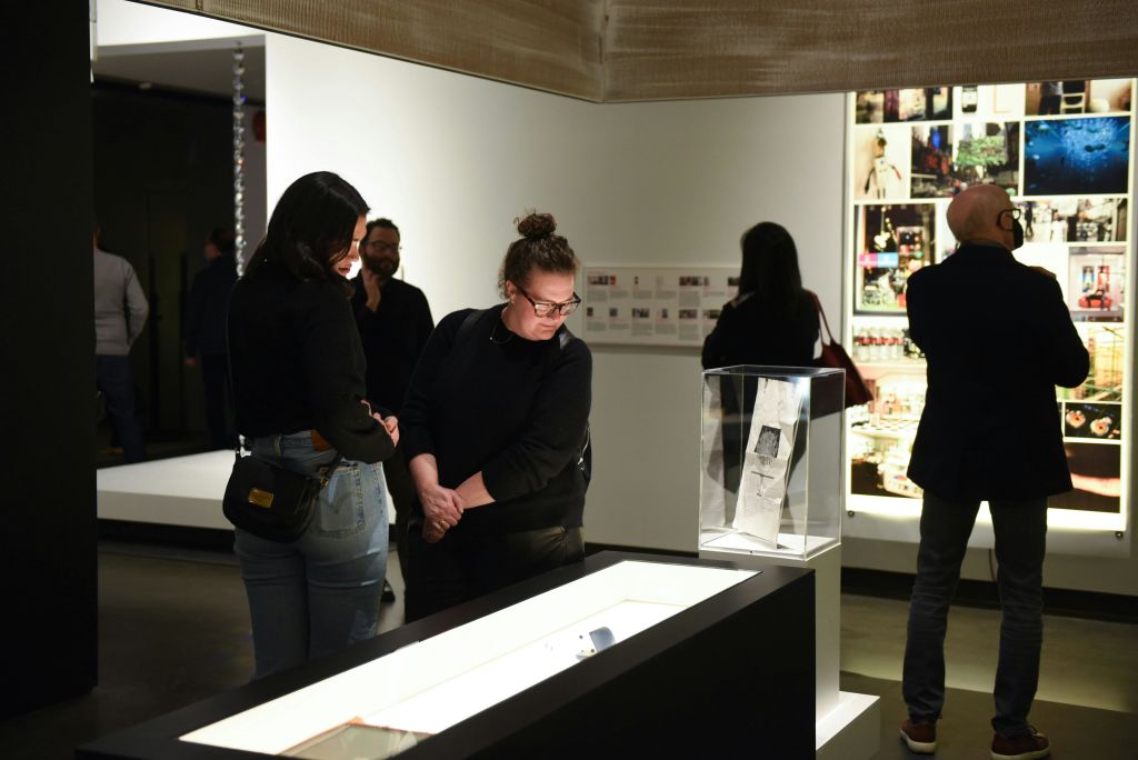 벽에 진열 케이스와 사진이 있는 조명이 희미한 박물관에서 전시물과 유물을 관찰하는 사람들.