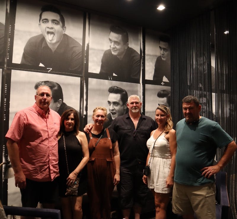 다양한 표정을 짓는 남자의 흑백 사진이 장식된 벽 앞에서 포즈를 취하는 여섯 사람.