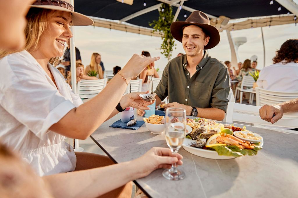 갈색 모자와 초록색 셔츠를 입은 남성이 해산물과 음료가 놓인 야외 테이블에 앉아 사람들에게 둘러싸인 채 미소를 짓고 있습니다.