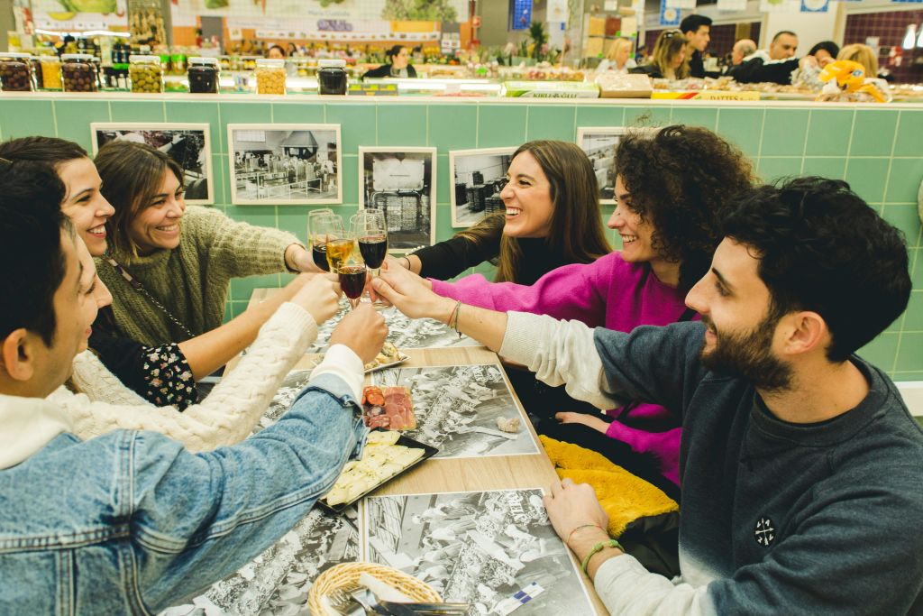 활기차고 밝은 실내 시장에서 음식과 함께 테이블 주변에서 음료를 마시며 건배하는 여섯 명의 친구.