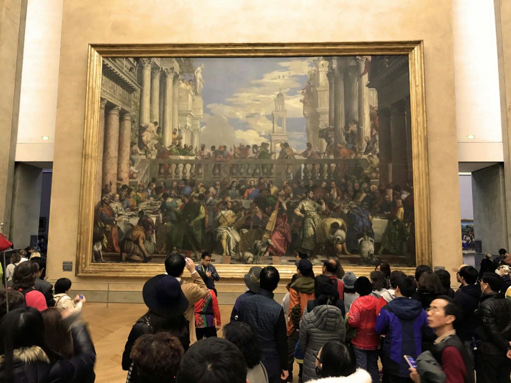 한 무리의 사람들이 박물관에 전시된 크고 섬세한 그림 앞에 모였습니다.