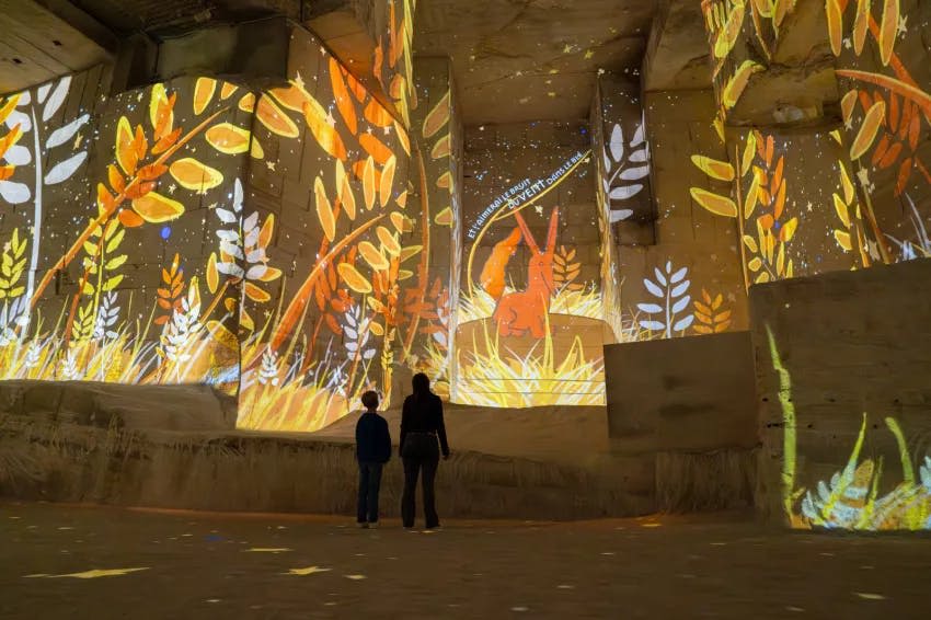 주황색과 노란색 나뭇잎 이미지와 토끼 같은 형상이 투영된 커다란 조명 공간에 두 사람이 서 있습니다.