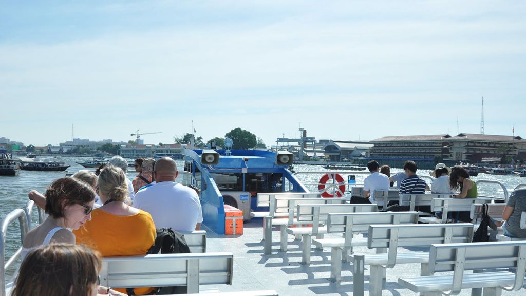 푸른 하늘 아래 강과 도시 풍경을 배경으로 한 보트에 벤치에 앉아 있는 사람들.
