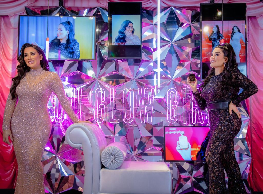 흰색 소파 옆에서 "You Glow Girl" 네온 사인과 그녀의 이미지가 표시된 스크린을 보며 포즈를 취하는 반짝이는 드레스를 입은 여성.