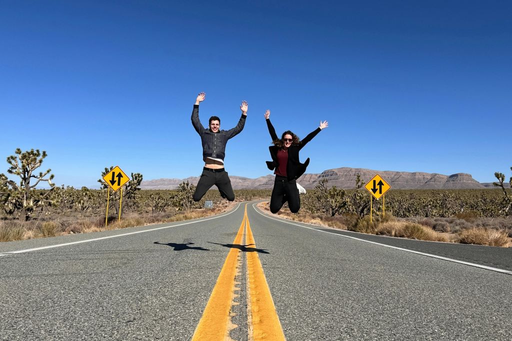맑고 푸른 하늘 아래 사막 지형과 교통 표지판으로 둘러싸인 텅 빈 도로에서 두 사람이 두 팔을 들고 점프합니다.