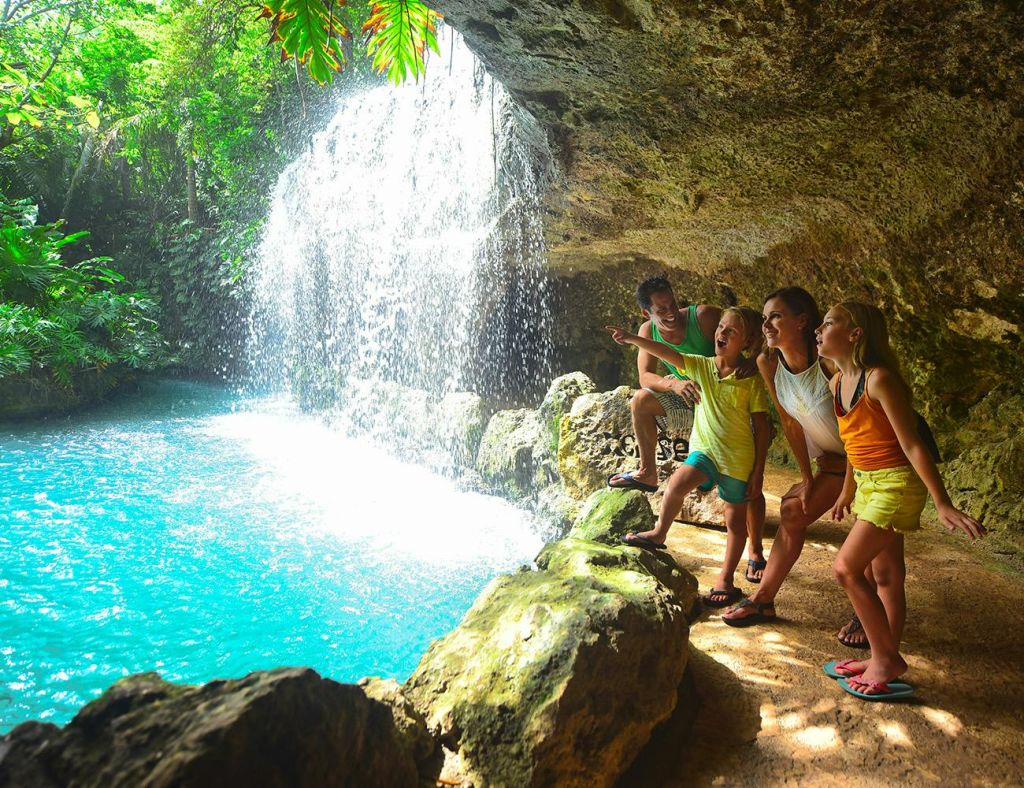 4인 가족이 바위 틈새에 서서 청록색 수영장으로 이어지는 폭포를 가리키며 미소를 짓고 있습니다.