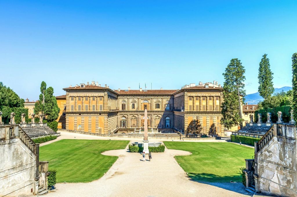 푸른 하늘 아래 푸른 잔디밭이 펼쳐진 대칭형 외관을 갖춘 크고 유서 깊은 건물입니다. 길을 걷는 사람들을 볼 수 있습니다.