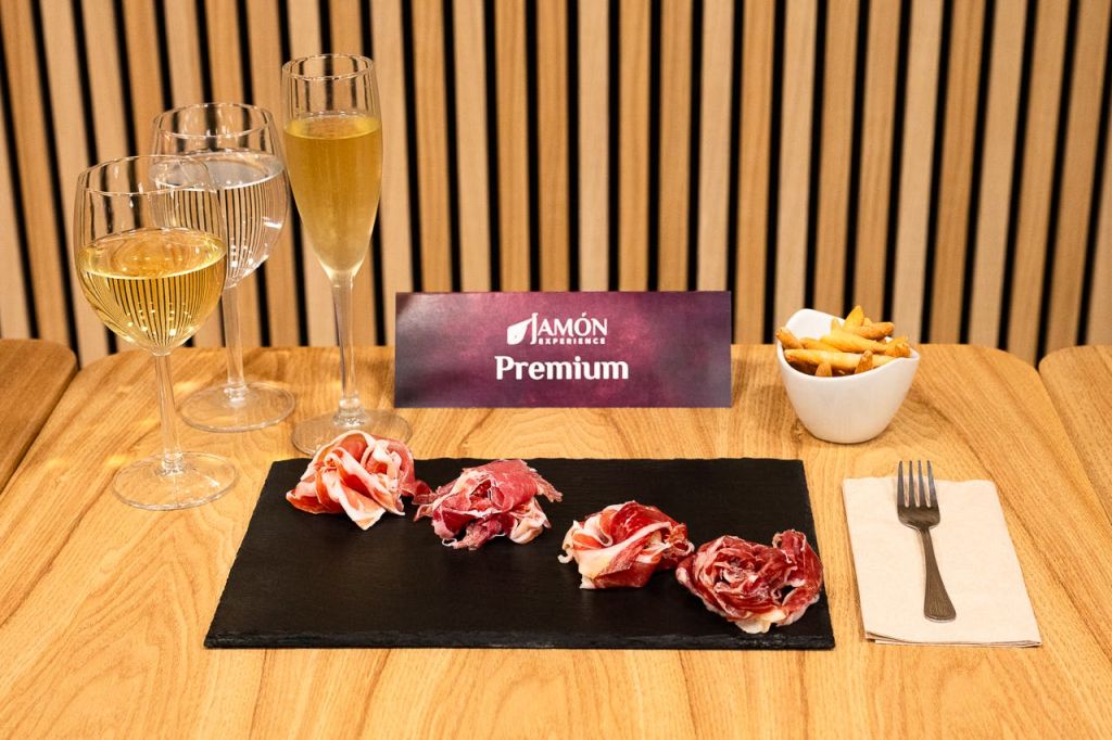 다양한 경화 고기, 스파클링 와인 2잔, 감자튀김 한 그릇, "Jamón Experience Premium" 표지판이 있는 슬레이트 접시.