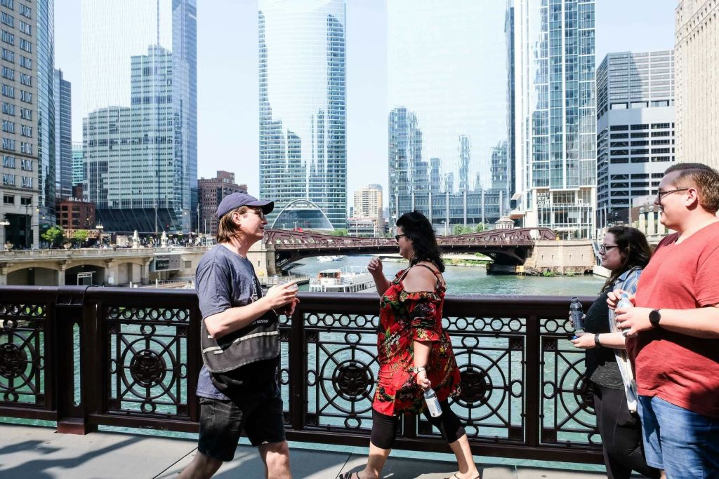 화창한 날 고층 빌딩과 투어 보트를 배경으로 강을 건너는 다리 위를 걷는 사람들.