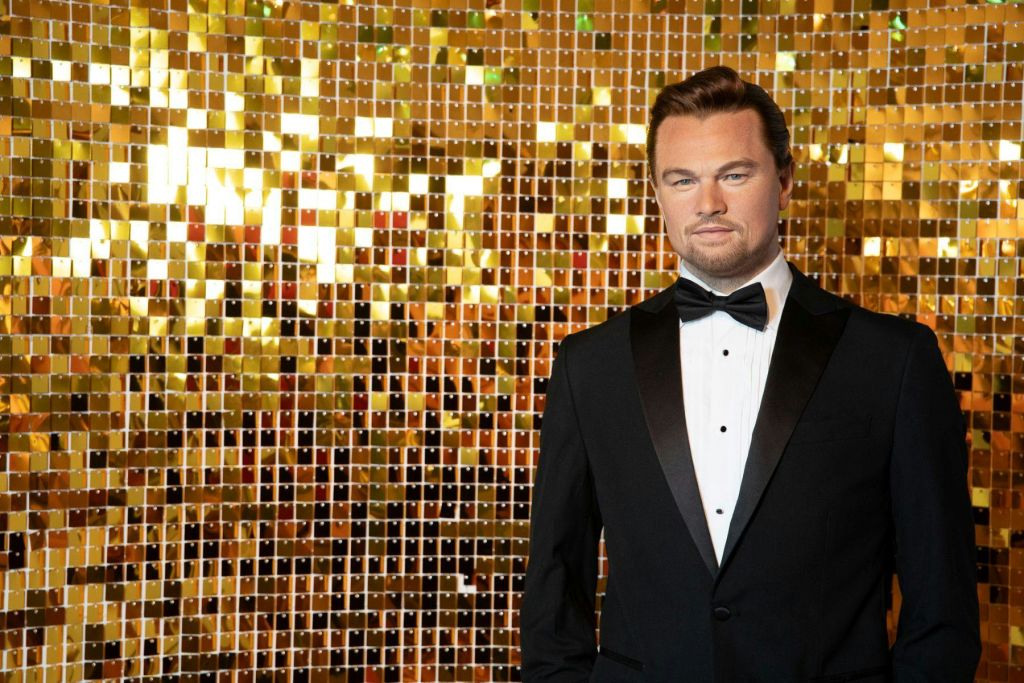 검은색 턱시도를 입은 남성이 중립적인 표정으로 골드 스팽글을 배경으로 서 있습니다.