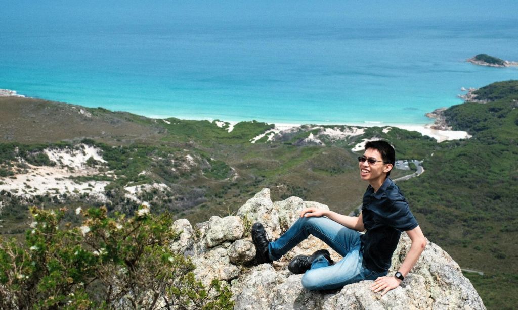 울창한 녹음과 청록색 바다를 배경으로 바위 절벽에 앉아 미소를 짓는 사람.
