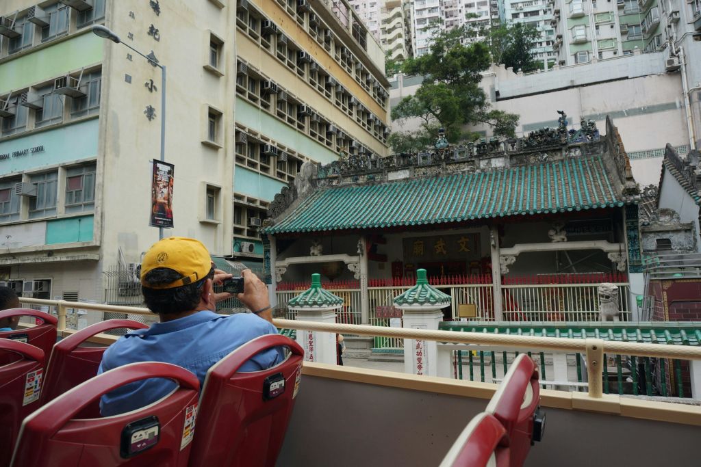 A person in a yellow cap takes a photo of a traditional Chinese temple from an open-top bus in an urban setting.