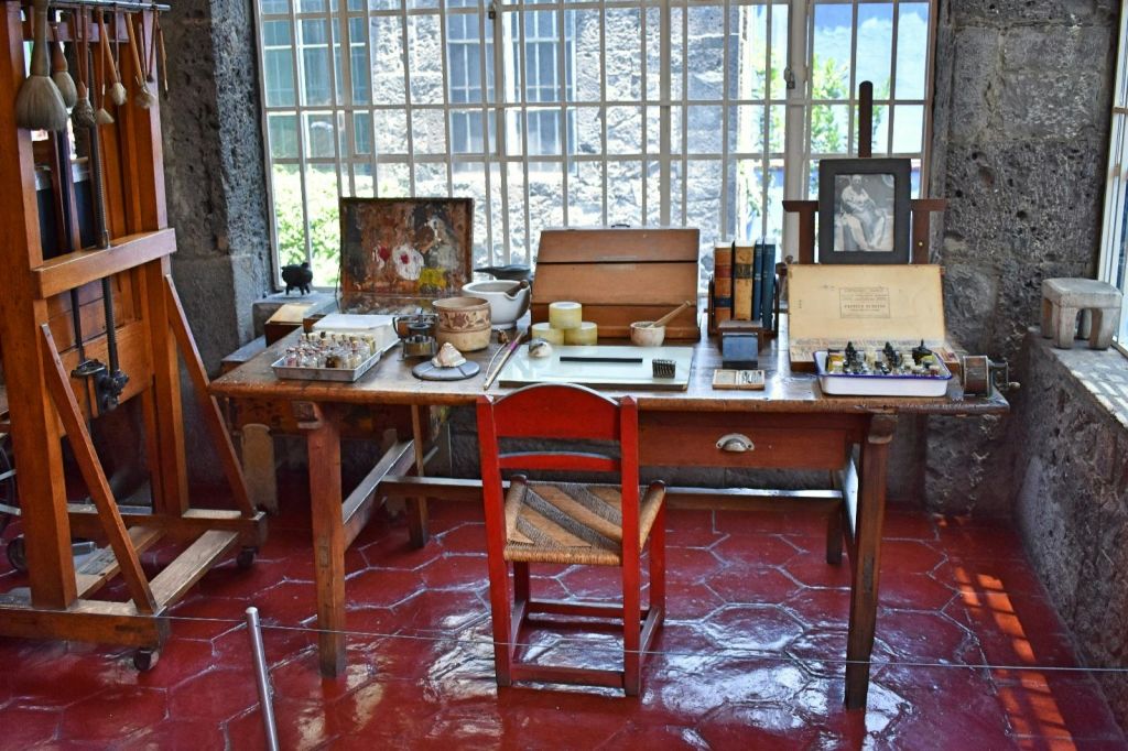 금속 막대가 있는 커다란 창문 앞에 페인트, 붓, 책, 예술품이 놓인 예술가용 책상이 있습니다.