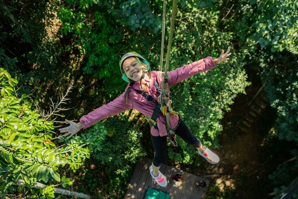 헬멧과 하네스를 착용한 사람이 숲을 가로질러 집라인을 펴고 미소를 지으며 팔을 바깥쪽으로 뻗고 있습니다.