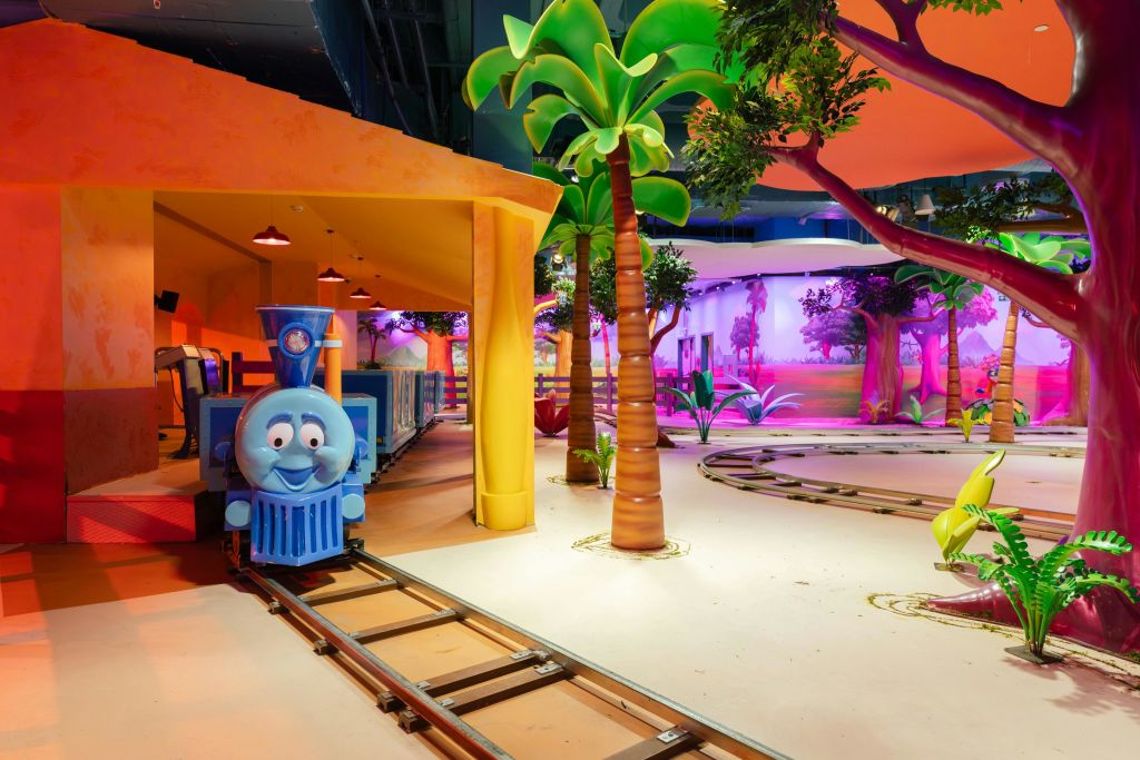 트랙, 야자수, 생동감 넘치는 조명 효과에 파란색 만화 기차가 있는 다채로운 실내 놀이 공간.