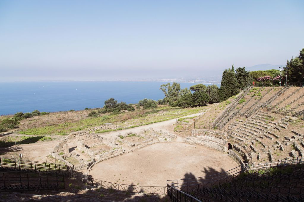 나무가 우거진 해안 풍경과 맑고 푸른 하늘이 내려다보이는 석조 좌석과 탁 트인 원형 무대가 있는 고대 원형극장.