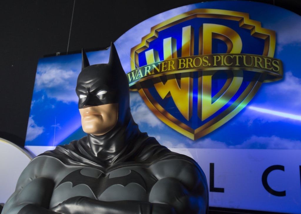 워너 브라더스 로고 앞에 팔짱을 끼고 하늘을 배경으로 한 배트맨 동상.