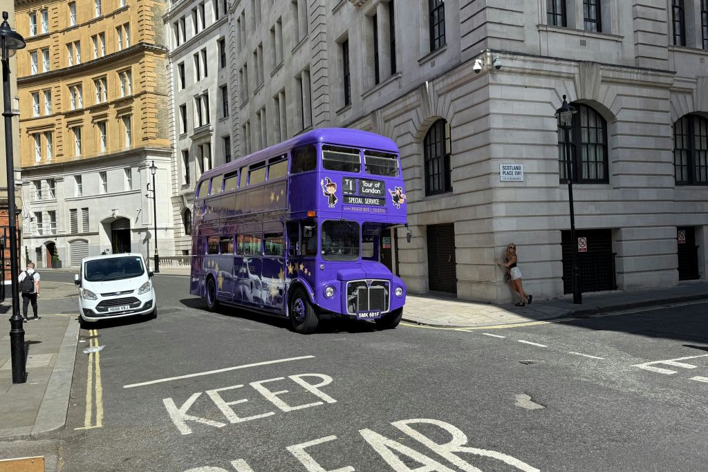 높은 건물 근처의 도시 거리에 있는 보라색 2층 버스입니다. 보도의 벽 옆에 사람이 서 있습니다.