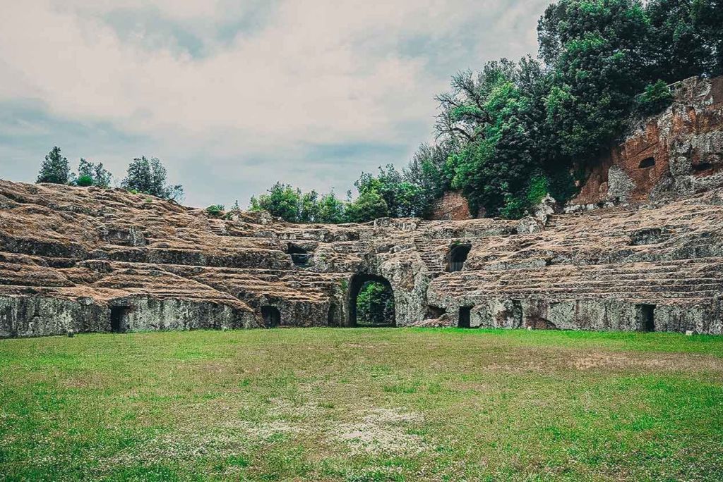 탁한 하늘 아래 잔디와 나무로 둘러싸인 석조 아치와 녹지가 있는 원형 극장의 고대 유적지.