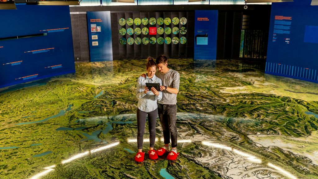 한 여성과 남성이 커다란 지도 바닥에 서서 흰색 십자가가 달린 빨간색 슬리퍼를 신고 태블릿을 보고 있습니다.