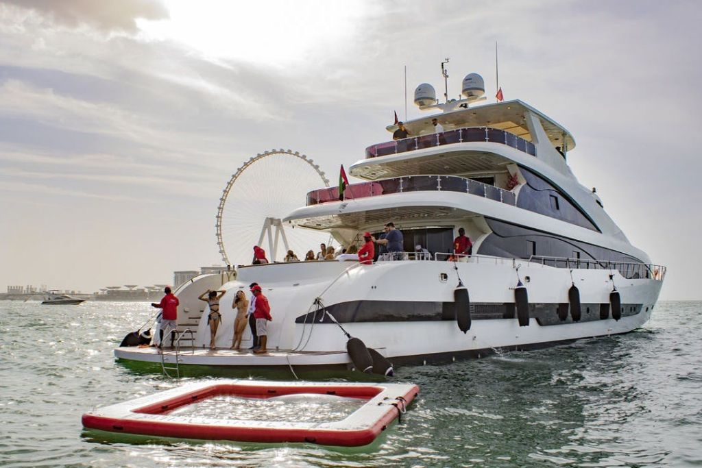 물웅덩이가 있는 멀티 데크 요트에서 수영복을 입은 사람들이 근처에 떠 있습니다. 대관람차가 배경에 보입니다.