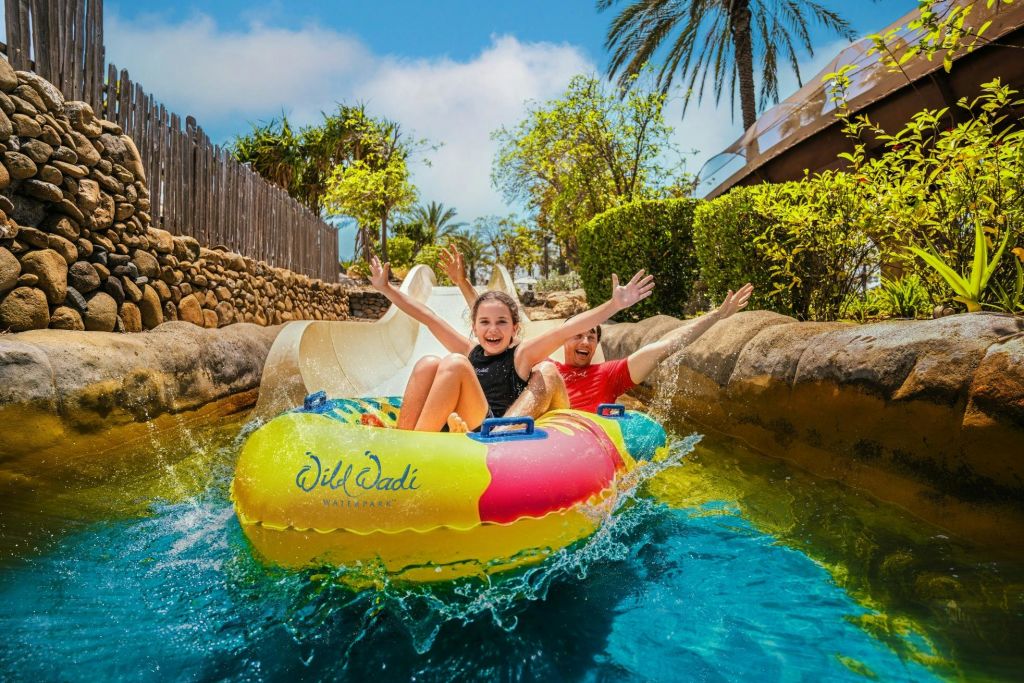 화초와 야자수로 둘러싸인 야외 공원에서 다채로운 색상의 팽창식 뗏목을 타고 미끄러져 내려오는 두 사람.