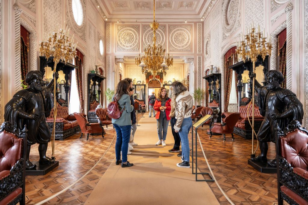 방문객들은 화려한 벽, 샹들리에, 빈티지 가구, 나무 바닥이 있는 호화로운 장식의 방에 서 있습니다.