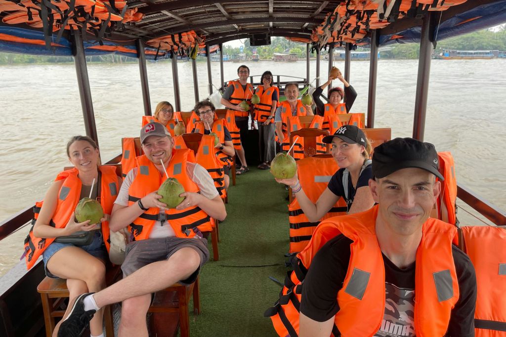 코코넛을 들고 있는 주황색 구명조끼를 입은 한 무리의 사람들이 구명조끼를 입은 채 보트에 앉아 있습니다.