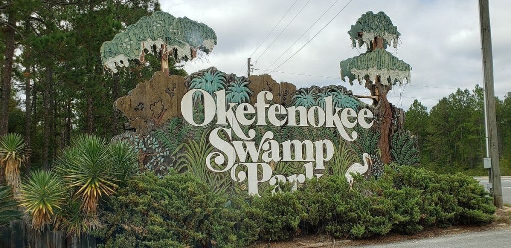 그림이 그려진 나무와 식물로 둘러싸인 "Okefenokee Swamp Park" 라는 표지판과 오른쪽에 새 그림이 있습니다.