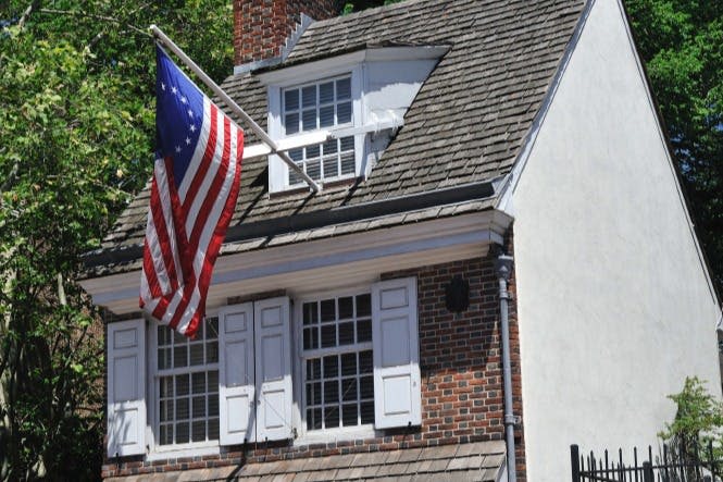흰색 창문 셔터와 지붕이 있는 벽돌 주택으로, 미국 국기가 전시되어 있으며 나무로 둘러싸여 있습니다.