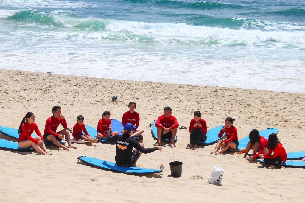 서핑보드가 달린 빨간 셔츠를 입은 한 무리의 사람들이 바다 근처의 모래 해변에 앉아 강사의 말을 듣고 있습니다.