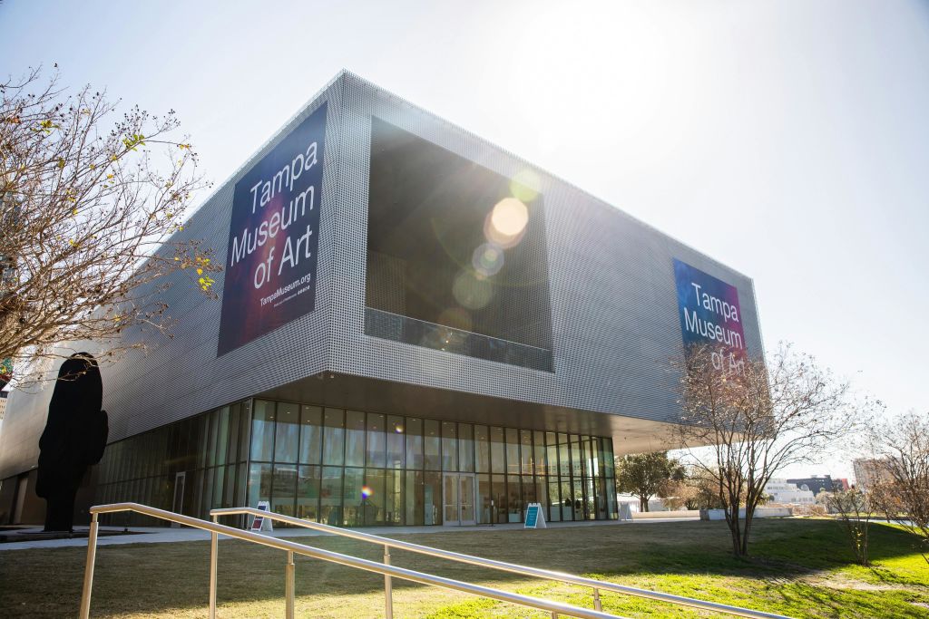 햇살이 내리쬐는 야외 공간으로 둘러싸인 유리 벽과 대형 배너가 있는 탬파 미술관의 현대적인 건물입니다.