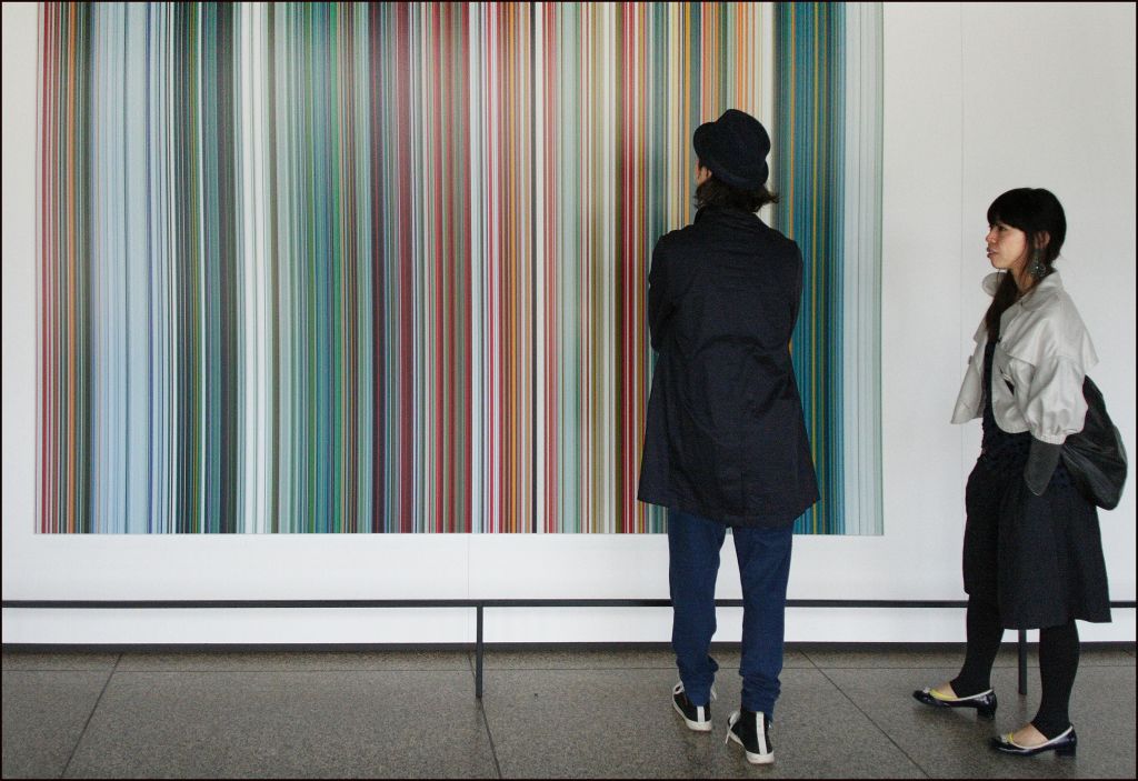 코트를 입은 두 사람이 갤러리를 배경으로 세로로 여러 가지 색상의 줄무늬가 있는 대형 작품을 관찰합니다.