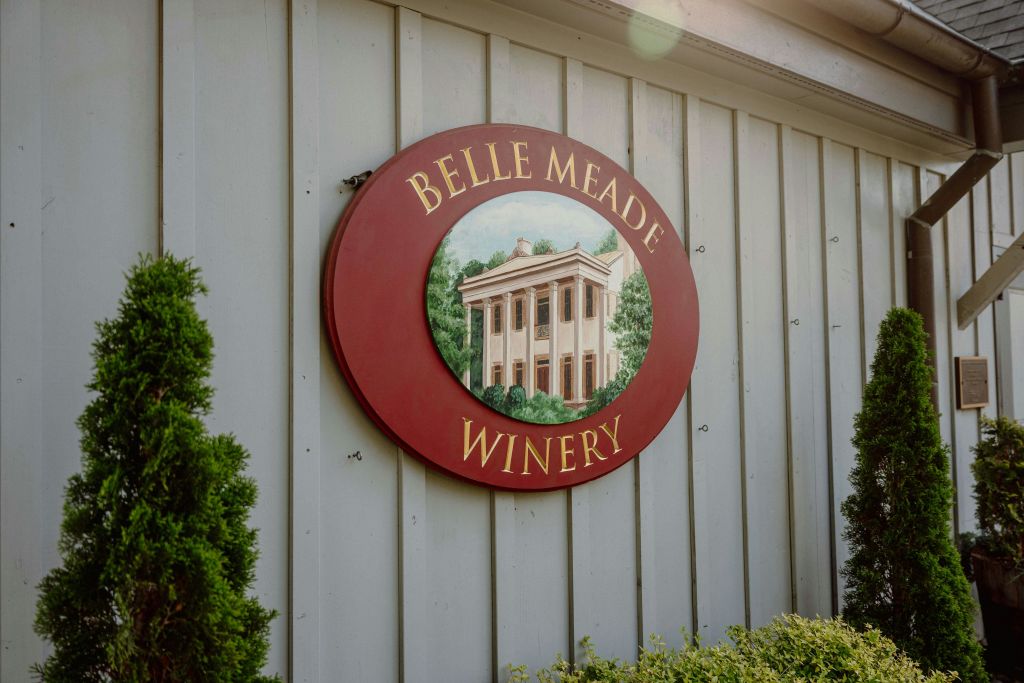 골드 레터링의 "Belle Meade Winery" 가 있는 둥근 빨간색 간판과 회색 나무 패널 벽에 장착된 건물의 그림.