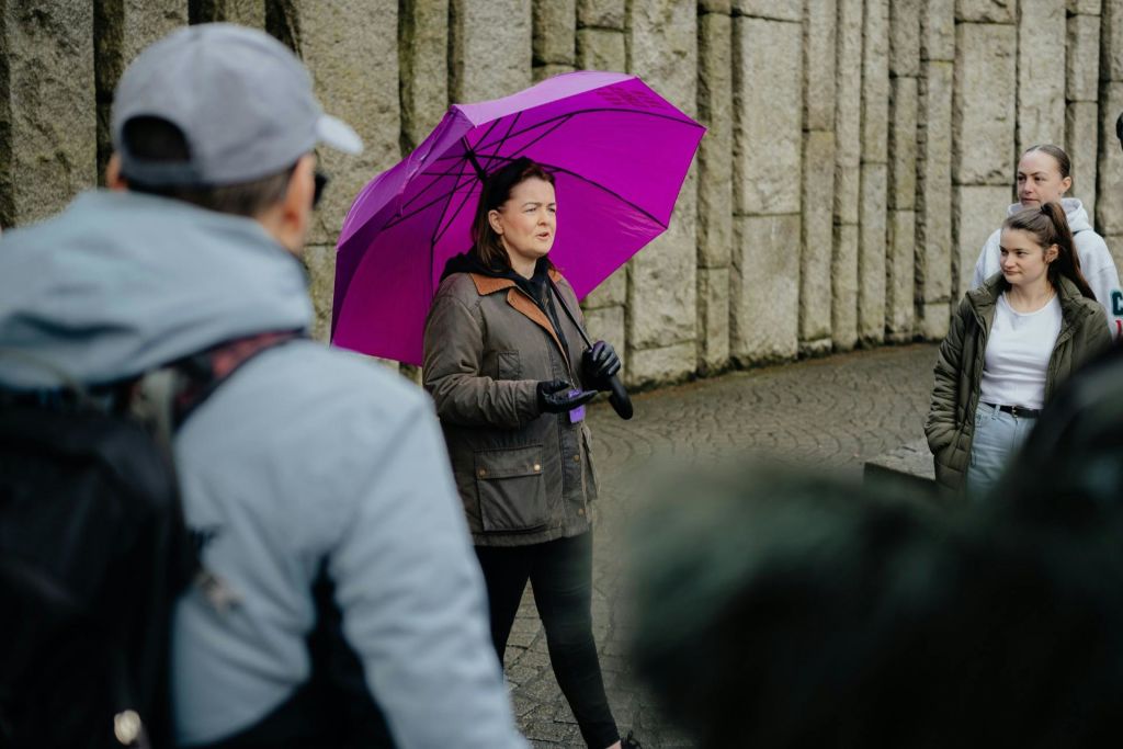 한 여성이 돌담 근처에서 단체와 대화하는 동안 보라색 우산을 들고 있습니다. 그룹은 전면에 부분적으로 표시됩니다.