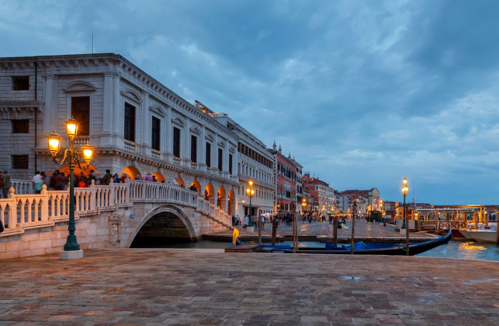 돌다리, 조명이 켜진 가로등, 유서 깊은 건물이 있는 베네치아 운하의 저녁 풍경. 사람들이 산책로를 따라 걷고 있습니다.