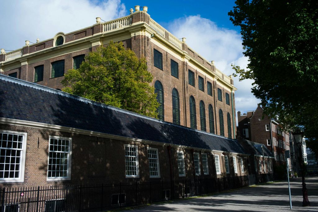 구름이 흩뿌려진 푸른 하늘 아래, 하부 구조에 인접한 커다란 아치형 창문이 있는 유서 깊은 벽돌 건물.