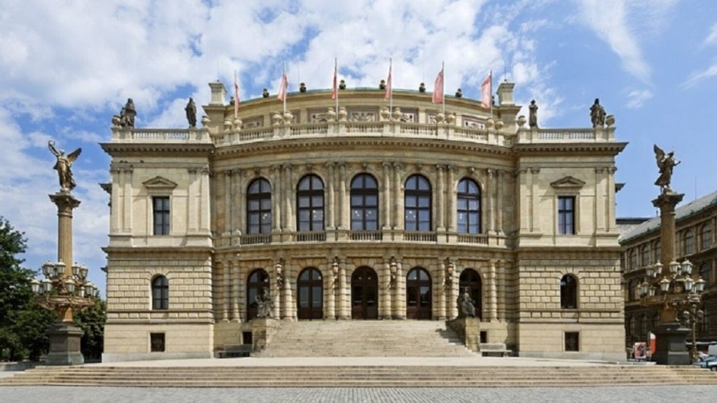 부분적으로 흐린 하늘을 배경으로 옥상에 아치형 창문, 조각상, 깃발이 있는 크고 화려한 건물.