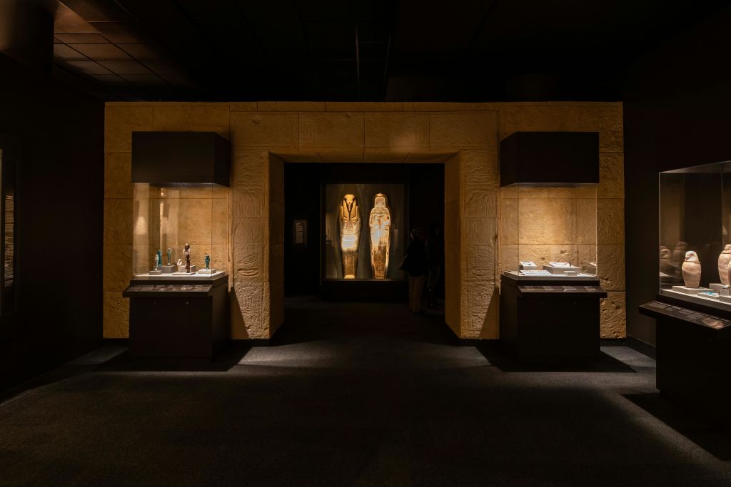 조명이 켜진 이집트 석관 두 개가 있는 희미한 조명의 박물관 전시관으로, 양쪽에 유물 케이스가 전시되어 있습니다.