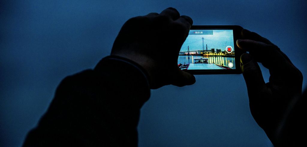 해질 무렵 불이 켜진 도크와 도시 풍경을 담은 스마트폰을 들고 있는 손.