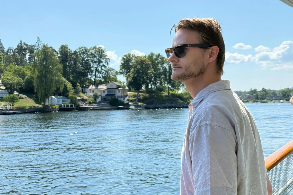 선글라스를 쓴 남자는 집과 나무, 부분적으로 흐린 하늘을 배경으로 배에서 물을 바라봅니다.