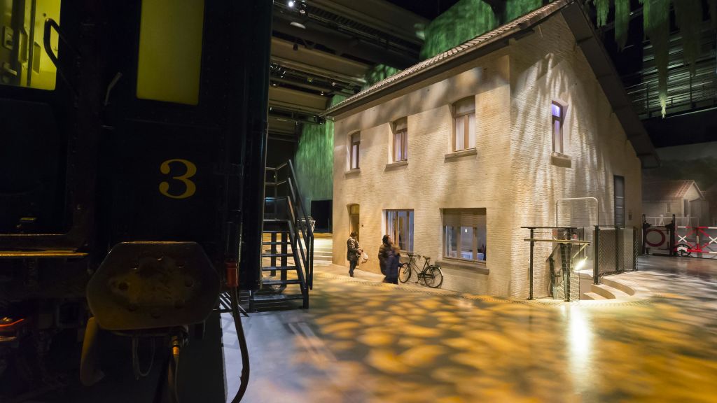 벽돌 건물 근처에서 사람들이 자전거를 끌고 대화를 나누고 있습니다. 그림자가 있는 계단과 녹색 벽이 배경에 있습니다.