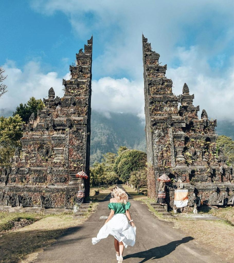 초록색 셔츠와 흰색 치마를 입은 사람이 산과 흐린 하늘을 배경으로 화려한 석문을 향해 걸어가고 있습니다.