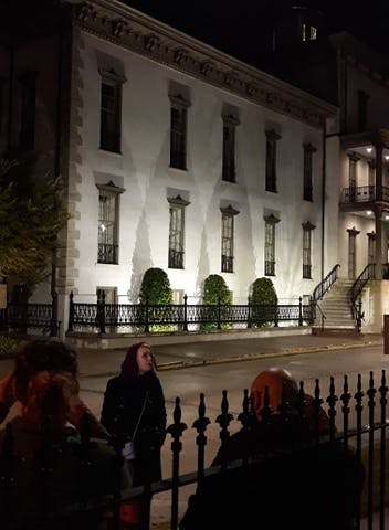 한 무리의 사람들이 밤에 보도에 서서 높은 창문과 철제 울타리가 있는 불이 켜진 역사적인 건물을 마주하고 있습니다.