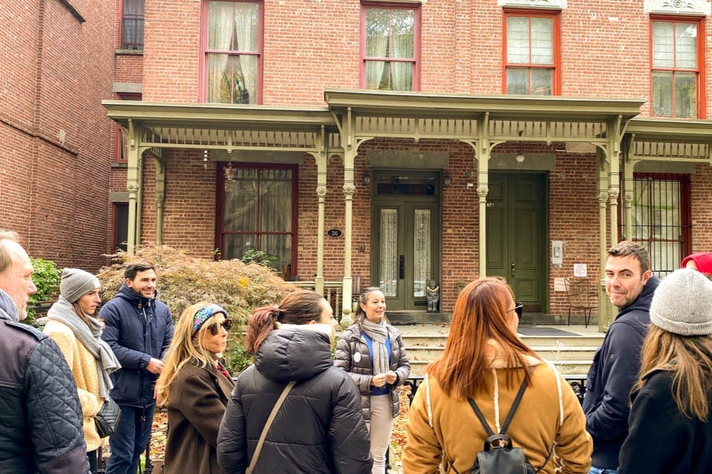 녹색 문과 커다란 창문이 있는 유서 깊은 벽돌 건물 앞에 한 무리의 사람들이 서서 대화를 나누고 미소를 짓고 있습니다.