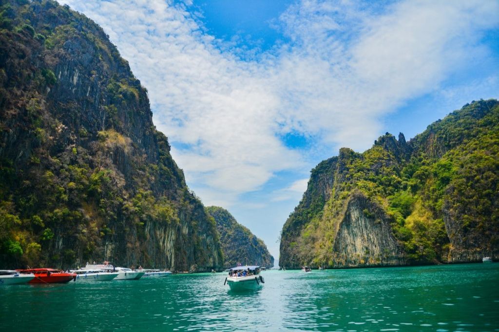 부분적으로 흐린 푸른 하늘 아래 녹색으로 덮인 우뚝 솟은 두 개의 절벽 사이를 항해하는 보트와 다른 보트를 배경으로 한 보트.