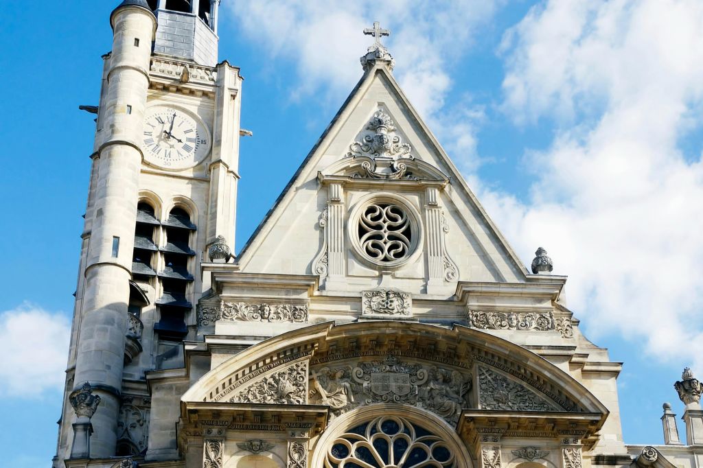 화려한 조각, 시계탑, 장미 창문, 구름이 거의 없는 푸른 하늘 아래를 가로지르는 고딕 양식의 교회 정면.